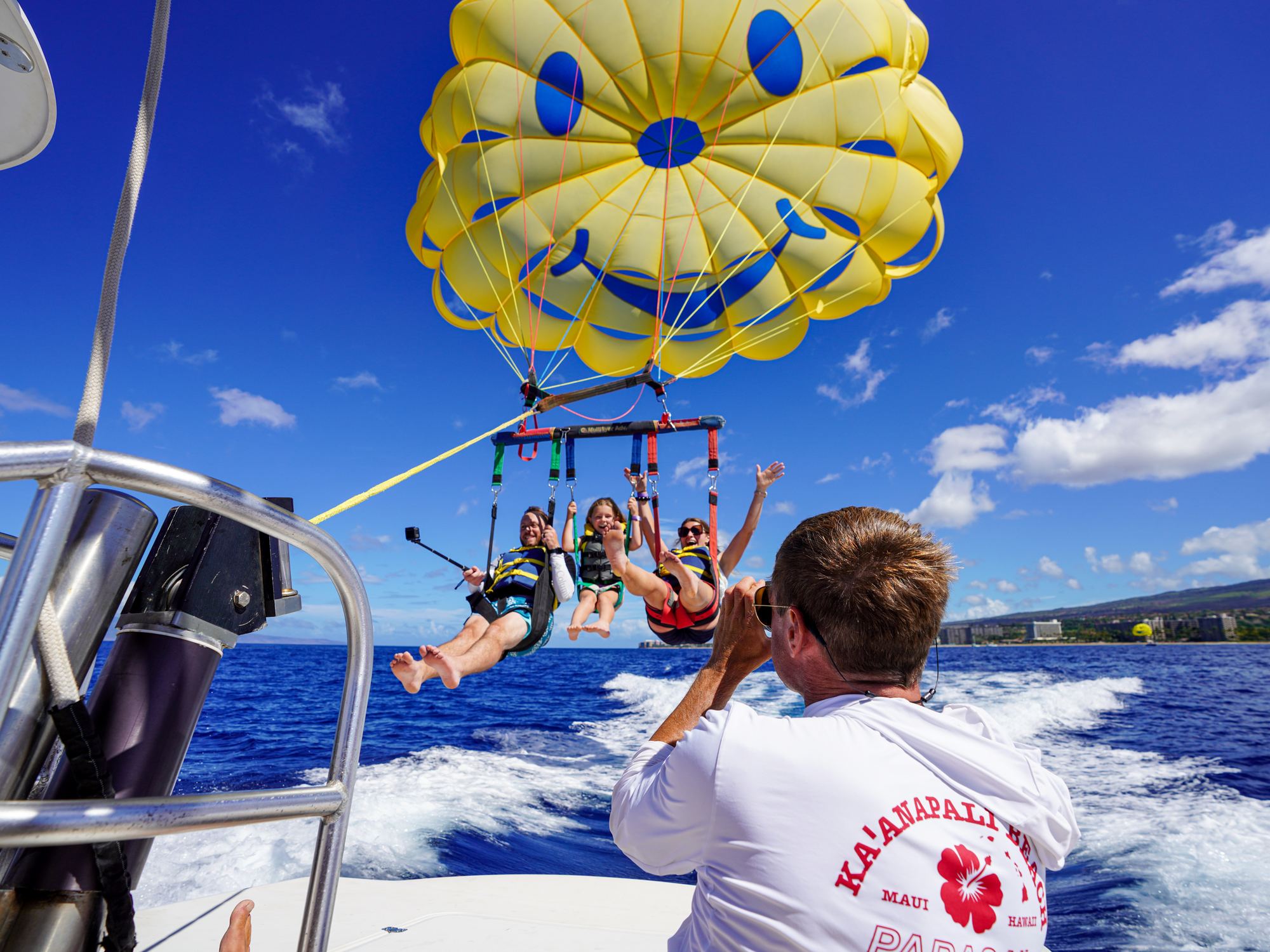 Three flyers airborne under the yellow smiley parasail, crew watching from the boat with the Kāʻanapali coastline behind